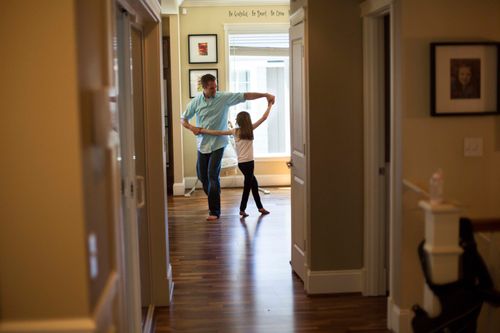 A father, wearing a blue shirt, and his daughter are seen from across the hall, dancing in their living room together.