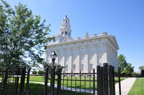 A side view of the Nauvoo Illinois Temple, with a black fence surrounding the grounds and a clear blue sky overhead.