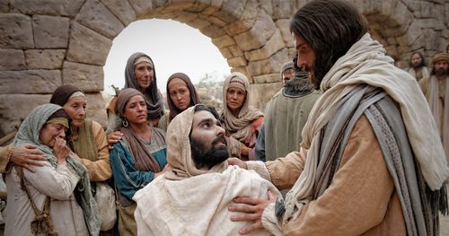 Młody mężczyzna owinięty w szaty pogrzebowe siedzący na noszach. Jezus trzyma rękę na jego ramieniu, a uczniowie Jezusa widoczni są w tle.