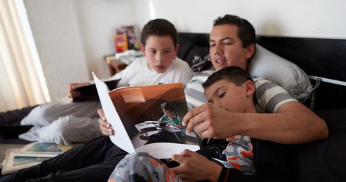 Father and two sons study together on a bed.