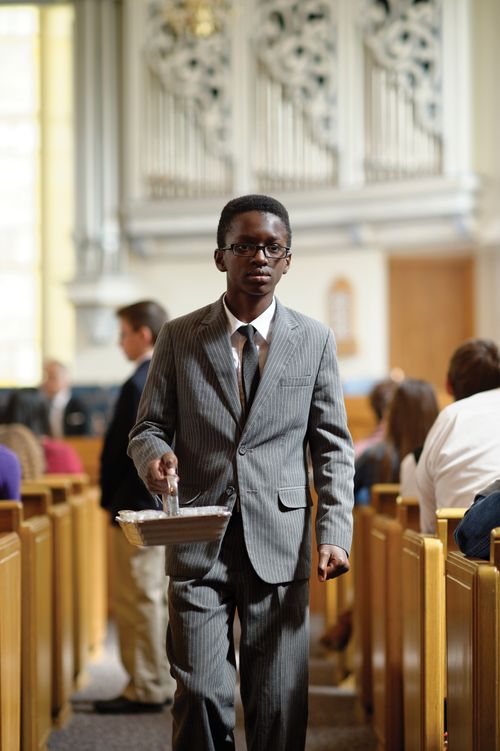 A young man in a suit carrying a sacrament tray along the aisles of a meetinghouse chapel.